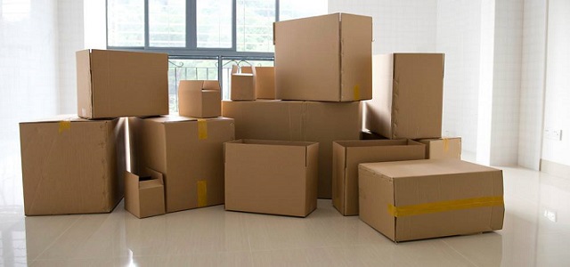 A pile of brown cardboard moving boxes in a large empty white room with windows.