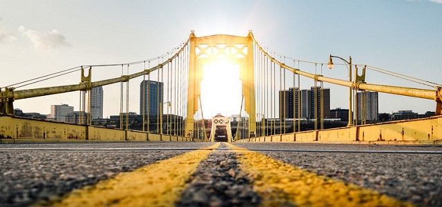 A blacktop road with two golden yellow traffic lines leading over a NYC bridge into the sunset.