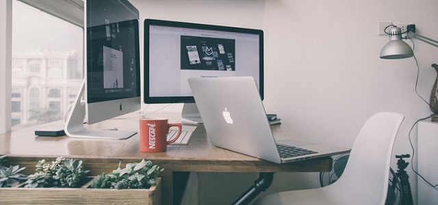 A brown wooden desk in front of a large window with two silver computer monitors and a silver laptop with a red coffee mug nearby.